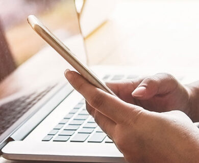 A persons hands around a phone checking news with a laptop in the background