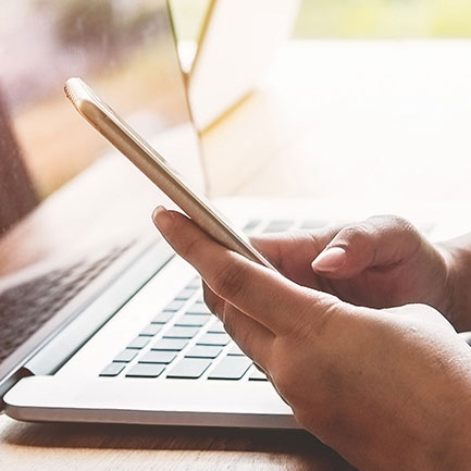A persons hands around a phone checking news with a laptop in the background
