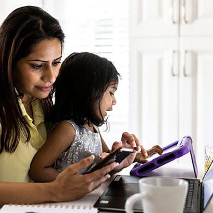 A woman scheduling online with her phone while a child in her arms uses a tablet to play a game